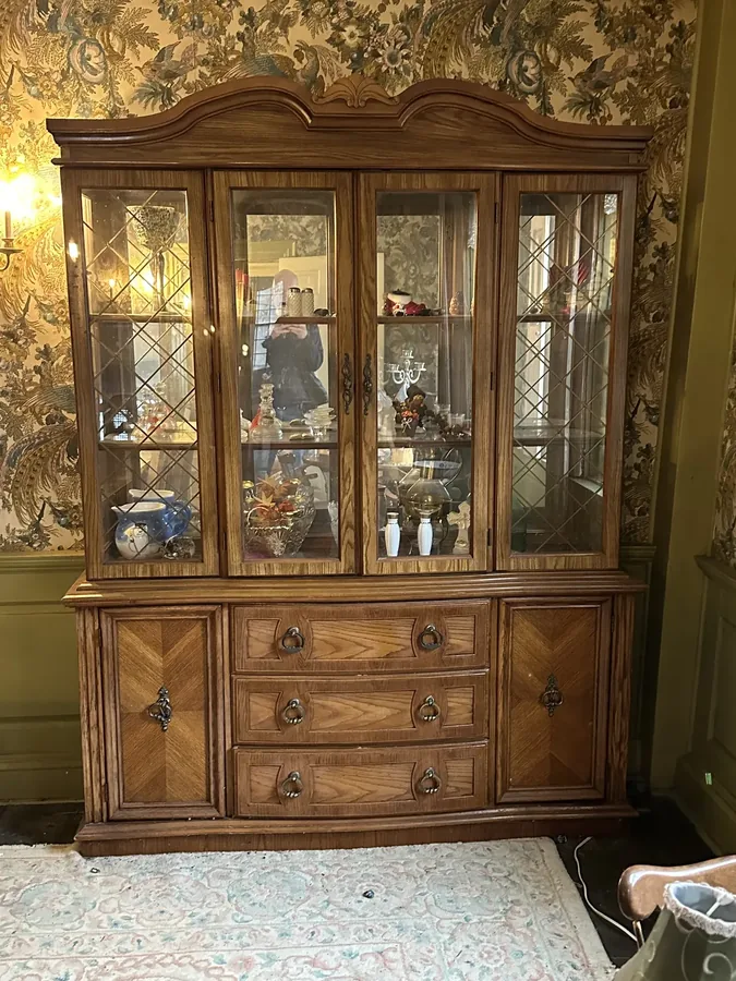 An antique china cabinet left behind, still holding glassware and dishes, against floral wallpaper