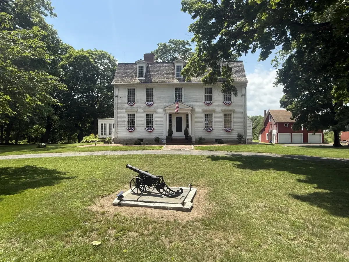 Front elevation of the General Henry Champion House in summer, with patriotic bunting, a cannon, and the barn visible